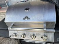 Photo showing Grill Master outdoor gas grill with four control knobs, stainless steel lid with temperature gauge, and side shelves. The exterior shows signs of dirt and grease buildup, indicating it needs cleaning.