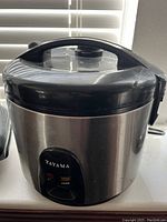 Front view of the Tayama metal rice cooker showing metal body, black plastic lid with steam vent, and front control panel with switch and indicator light.