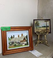 Wider view showing both framed art pieces with the wooden framed 3D house on the left and smaller landscape painting on metal stand on the right