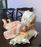 Side angle of porcelain baby doll posed crawling on table, showing peach dress, bonnet, and holding a lace-trimmed cushion, good detail of doll's face and hands.
