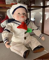 Porcelain baby doll dressed in white sailor suit with navy accents and red neckerchief sitting on shelf.