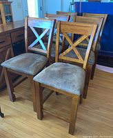 Front and side view of six wooden bar chairs with cross back design and gray upholstered seats arranged together on hardwood floor.