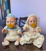 Two baby dolls seated on wooden chairs wearing vintage handmade crocheted outfits with yellow pacifiers in their mouths.