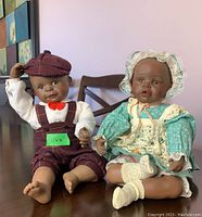 Two Yolanda Bello porcelain dolls, boy and girl, seated on a dark wooden table against a purple wall.