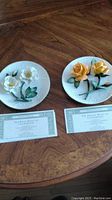 Pair of 3D Capodimonte floral plates with certificates on wood table.