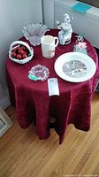 Photo showing entire lot on table with burgundy cloth: Lenox glass dish, two Schmidt musical items, ashtray, ceramic basket with cherries, glass floral figure