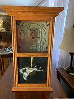 Front view of wooden mantel clock with clock face showing green numbers and duck silhouettes. Lower glass panel shows flying duck and pendulum.