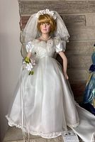 Front view of porcelain bridal doll in white gown, veil, and holding bouquet placed against a stone background.