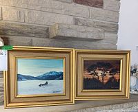 Two framed paintings on a stone mantel showing a snowy landscape with sled dogs on the left and a tree silhouette sunset/sunrise scene on the right.