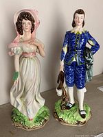 Front view of two porcelain figures, female in pink bonnet and male in blue costume, standing on green and brown bases