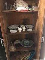 Shelf showing white ceramic plates and bowls, creamers, small square plates, folded lace doilies and tablecloths above, and various items below including glass cloche.