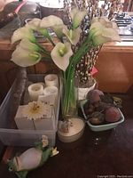 Photo of white artificial calla lilies in glass vase, white pillar candles in box, faux fruit bowl, and a small frog figurine on table.