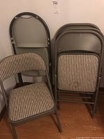 Six folding chairs, five upholstered fabric folding chairs with metal frames in beige stripes, one gray molded plastic folding chair, stacked and arranged near a wall on wood floor.