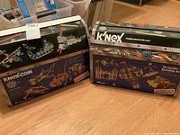 Two original K'NEX COM boxes each containing 700-piece building sets showing model ideas on the box, unopened plastic bags inside but boxes opened.