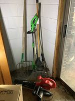 Overview photo of the lot showing Poulan electric chainsaw, Homelite blower, Expert weed eater, metal rake, and pitch fork leaning against a wall in basement.