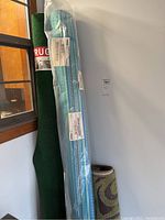 Rolled green Foss Floors indoor/outdoor area rug leaning against a wall with two other rugs rolled beside it.
