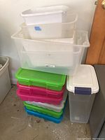 Photo showing a stack of clear plastic bins without lids on top, colorful plastic craft bins with lids underneath, a tall translucent container with lid on the side, and part of a white trashcan visible on the left.