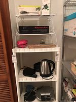 Plastic shelving unit with various small kitchen appliances and tools including KitchenAid spiralizer attachment box, herb tools, pink pizzelle iron, scale, coffee pot, kettle, and bunt maker.