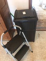 Two-step stool with metal frame and black steps placed beside the Suncast deck trash can indoors on printed tile floor near wooden door and cabinet.