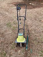 Photo showing front view of Sun Joe electric garden tiller with black handles and green body alongside the spade and garden claw on grass.