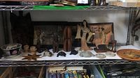 Wide view shelf display showing a variety of wooden and stone figurines, framed art, and bowls for inventory and content verification.