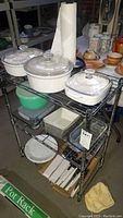 Three white Corningware ceramic baking dishes with glass lids on top shelf of metal pot rack, green lidded plastic containers and glass dishes below, tins and plates on lower shelves.
