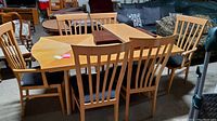 Dining table set featuring a rectangular light wood table with six matching chairs with upholstered seats, set inside a warehouse environment.