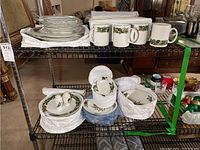 Overall view of multiple shelves displaying white ceramic dinnerware with holly garland decoration including stacked plates, mugs, bowls, and serving pieces.