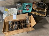 Overview of mid-century cocktail glasses in cardboard box, several decorative trays, 1939 World's Fair commemorative glasses, and Coca Cola straw dispenser on metal shelf.