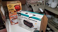 Boxed West Bend 4 Quart Slow Cooker Model 4000 in red and Proctor Silex 4 Quart Slow Cooker in white with orange accents stacked with Thermos Portable Gas Grill box alongside.
