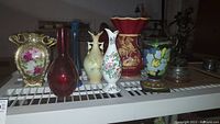 Full view of seven assorted vases and a figurine on white metal shelf, showing variety in styles and materials