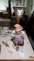 Full view of the vintage ceramic child figurine table lamp on a table, showing the figurine holding a basket and the ornate lamp shade above it.