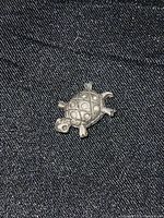 Close-up photo of silver-colored turtle bead showing detailed shell pattern and limb shapes with loop on top for stringing.