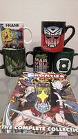 Front view of the five coffee mugs stacked and the Marvel Frontier graphic novel placed in front showing cover art