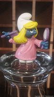 Photo showing vintage Smurfette figurine holding a pink brush and purple mirror, standing on a glass surface.