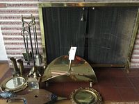 Photo showing brass fireplace screen, brass fireplace tools with stand, brass fire bellows, small round mirrors, brass ducks figurines, and a brass wood holder on tiled floor.