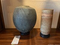 Two ceramic pottery pieces on teak display stands shown on a wooden surface with a lot tag for size indication.