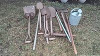 Photo showing six garden shovels laid out on grass, varying blade shapes and handle types visible, with metal watering can to the right.