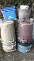 Full view of eight bins including two blue recycle bins, beige, brown, green, and grey waste bins stacked outdoors on gravel with rocks behind