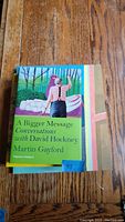 Top view of 'A Bigger Message: Conversations with David Hockney' book with colorful cover showing man with outstretched arm among a natural setting.