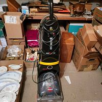 Full view of Bissell carpet cleaner among packed boxes and household items showing upright black and yellow cleaner with clear front tank.