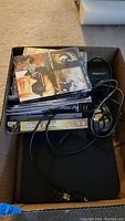 Photo of black DVD player with power and USB cables on top of a cardboard box with DVDs and cases