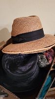 Tan woven straw hat with black band visible on top of other hats in a box.