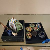 Various small decorative items and pill boxes displayed on black trays inside a glass cabinet, including a porcelain teapot-shaped container, a figurine, colorful floral and animal patterned pill boxes, and round containers.