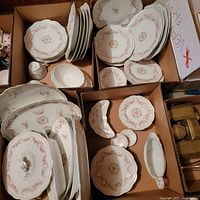 Wide angle photo showing multiple boxes filled with porcelain plates, serving platters, bowls, and a covered dish, all decorated with a pink floral pattern and gold trim.