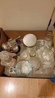 Box filled with various glass kitchenware including measuring cup, coffee press, jar with lid, and glass containers