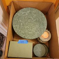 Top view of the lot in a cardboard box showing a green stone cake stand, matching green stone coasters, a small jar containing ceramic pie weights, and a sealed green box likely containing recipes.
