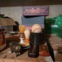 Photo showing all trinkets on a wooden surface including two shaving brushes, metal piggy bank shaped like a pig, a baseball inside a cylinder leather holder, two decorative boxes, two small boxes, and a long metal tool.