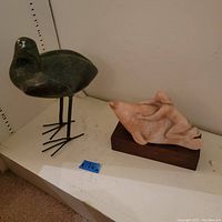 Photo showing both sculptures together on a white surface: dark green stone bird with metal legs and metal around eye, and pale stone woman on sea creature mounted on wood base.