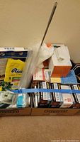 Photo showing assorted household supplies including tissue boxes, plastic gloves, disposable cups, cleaning duster, and packaged food item in boxes